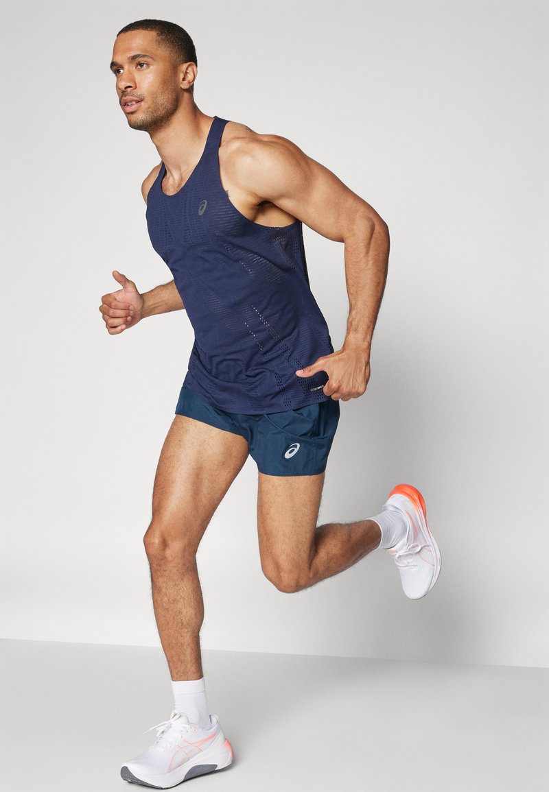 Marineblauer Sporttanktop mit Mesh-Struktur, kombiniert mit marineblauen Shorts. Weiße Laufschuhe mit orangefarbenen Akzenten, Modell in Bewegung vor einem hellen Hintergrund.