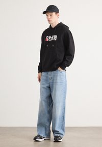 Svart hettegenser med hvit og rød logo, lyseblå baggy jeans og svart caps. Svarte og hvite joggesko fullfører antrekket.