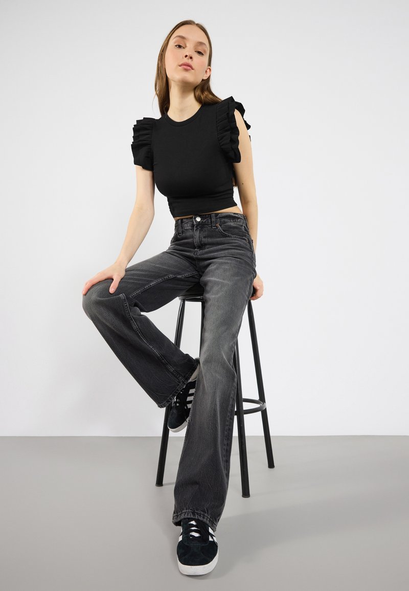 Černý crop top s volánovými rukávy v kombinaci s černými džínami s vysokým pasem a odřelým vzhledem. Model sedí na černém stoličce a má na sobě tmavé tenisky.
