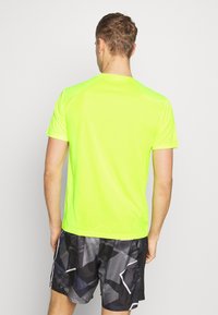 Fluorescerende gul kortærmet træningstrøje med en glat tekstur, båret med sorte shorts med et geometrisk mønster.