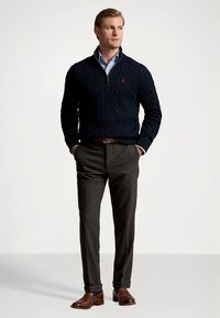 Granatowy sweter w splot żakardowy, z zamkiem przy kołnierzu, noszony na jasnoniebieskiej koszuli; szare, dopasowane spodnie; brązowe skórzane buty; minimalne elementy z logo.