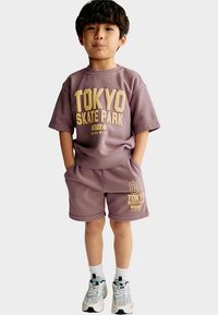 Dítě v růžovofialovém tričku s žlutým nápisem "TOKYO SKATE PARK", v odpovídajících šortkách a bílých ponožkách s teniskami.