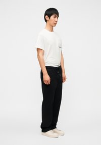 T-shirt en coton blanc avec un détail graphique sur la poche, associé à un pantalon de jogging noir et des baskets de couleur claire. Vue de profil sur un fond uni.