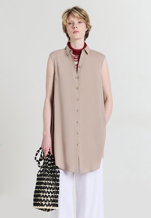 Giovane che indossa una tunica beige senza maniche con bottoni e pantaloni bianchi, tiene una borsa intrecciata bianco e nero e indossa una collana rossa a più strati.