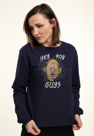 Kvinde iført marineblå sweatshirt med grafik af en skaldet mands ansigt og tekst 'Hey you guys', kigger ned og holder i kanten af sweatshirten.
