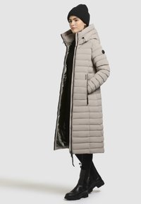 Lunga giacca ecopelliccia grigia chiara con cappuccio, dotata di chiusura a zip, tasche laterali e polsini a coste; abbinata a stivali neri e un berretto.