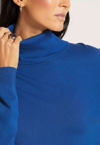 Blauer Rollkragenpullover mit gerippter Textur, hochgeschlossen und mit langen Ärmeln. Metallischer Ring und Ohrringe sorgen für Kontrast.