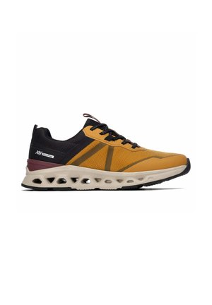 Zapatilla deportiva baja en amarillo mostaza y negro con cordones negros, suela blanca acolchada con orificios de ventilación y detalle de marca en el talón.