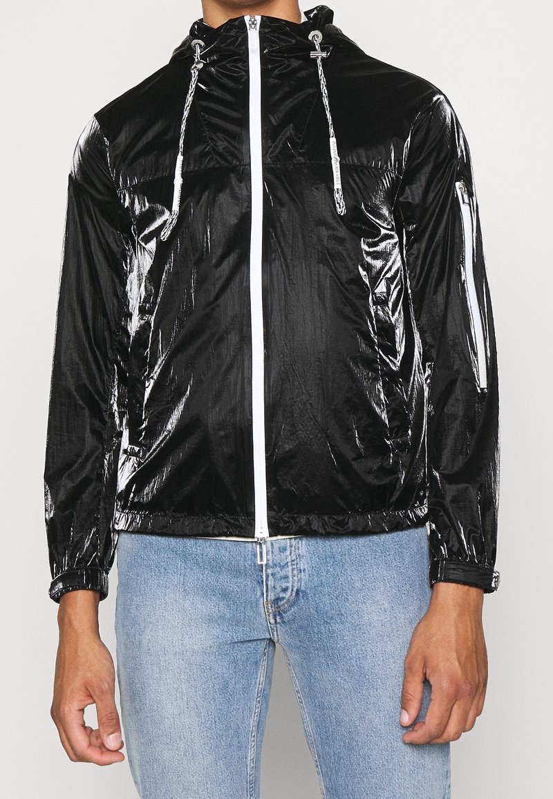Svart glansig jacka med huva, dragkedjestängning och elastiska ärmmuddar; har reflexdetaljer och en framficka med dragkedja.