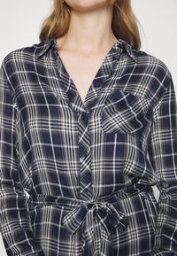 Femme portant une chemise à carreaux bleu marine, blanc et noir, boutonnée, avec une taille nouée et une poche poitrine, vue du cou jusqu'aux hanches.