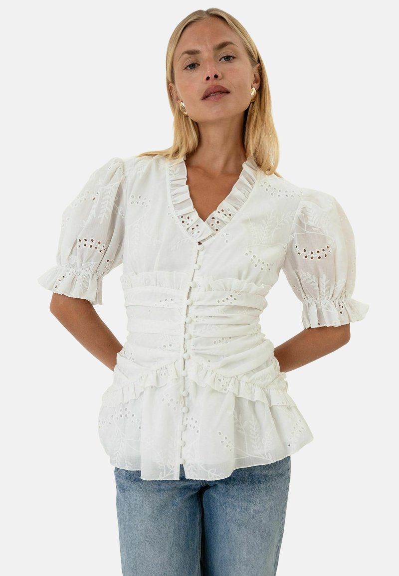 Femme portant un chemisier blanc à broderie anglaise avec manches bouffantes, bord à volants, et jean bleu, debout les mains derrière le dos.