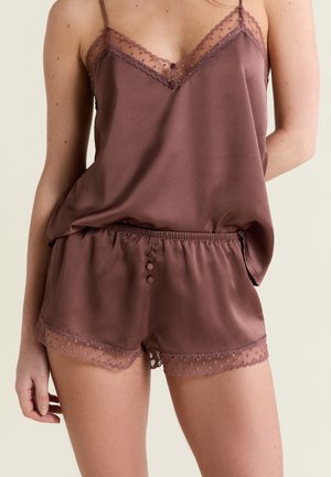 Frau trägt mauvefarbenes Satinkleidchen und passende Shorts mit Spitzenbesatz und Knopfdetails vor hellem Hintergrund.