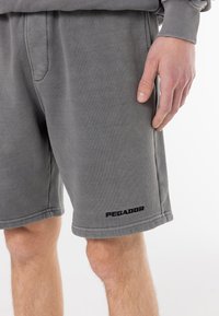 Mann iført grå knelange shorts med svart "PEGADOR"-logo på venstre ben og matchende grå genser, står foran hvit bakgrunn.