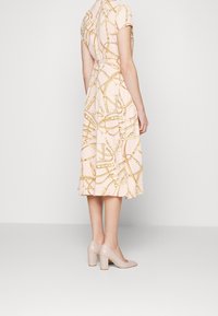 Vestido de color rosa claro con un patrón de cadenas doradas, mangas cortas, un dobladillo acampanado y un corpiño ajustado. Combinado con zapatos de tacón cuadrado en color nude.