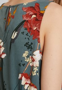 Top bez rękawów w wzór kwiatowy na tkaninie w kolorze teal. Z czerwonymi i białymi kwiatami, dekoltem z wycięciem na plecach oraz miękką fakturą.