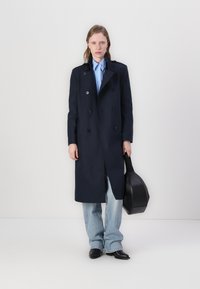 Palton bleumarin, cu două rânduri de nasturi mari, asortat cu o cămașă bleu deschis și blugi lejeri din denim deschis. Ține o poșetă neagră.