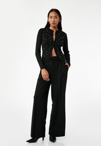 Cardigan nero a coste con piccole perle sparse, chiuso con bottoni, abbinato a pantaloni neri a vita alta e décolleté neri con punta.