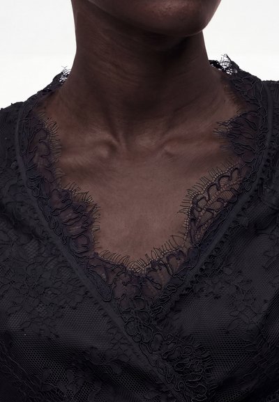Κοντινό του λαιμού και του στήθους ενός ατόμου που φοράει μαύρο δαντελωτό ένδυμα με περίπλοκα άνθη και κυματιστά άκρα.