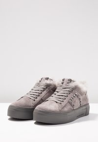 Baskets en suède gris avec doublure en fourrure, accents cloutés sur les côtés, semelles épaisses en caoutchouc et lacets, présentées sur une surface blanche.