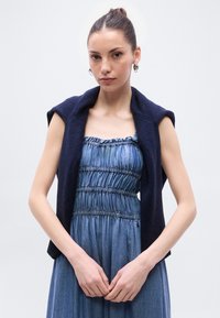 Ναυτικό μπλε πλεκτό γιλέκο συνδυασμένο με φόρεμα από τζιν με μαζεμένη υφή και ρυθμιζόμενους ιμάντες· και τα δύο ρούχα έχουν άνετη εφαρμογή.