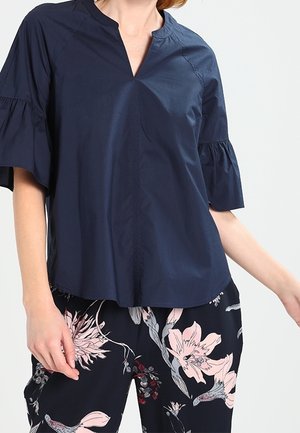 Femme portant un chemisier bleu marine à manches courtes avec des manches à volants et un pantalon à motif floral avec des fleurs roses et blanches.