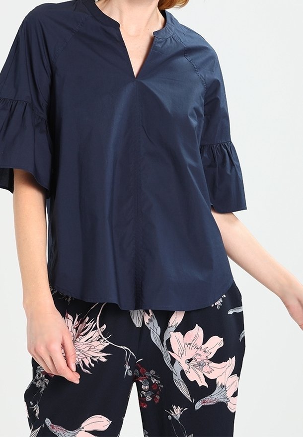 Femme portant un chemisier bleu marine à manches courtes avec des manches à volants et un pantalon à motif floral avec des fleurs roses et blanches.