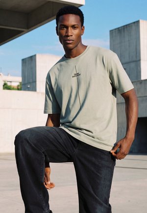 Fiatal férfi világosszürke "Think Different." feliratú pólóban és fekete nadrágban, szabadtéren, városi beton környezetben pózol, tiszta ég alatt.