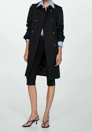 Femme portant un trench noir sur une chemise à rayures bleues, un pantalon noir raccourci et des sandales à talons noires, debout devant un fond uni.