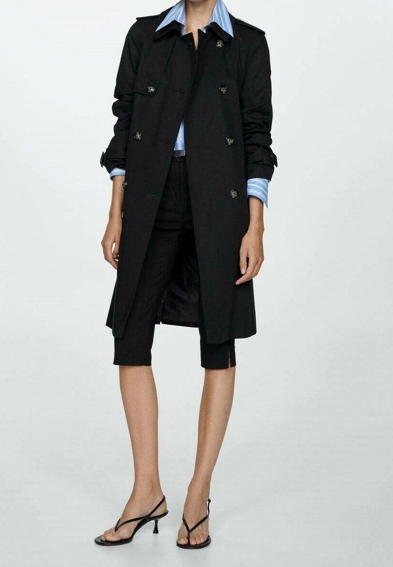 Femme portant un trench noir sur une chemise à rayures bleues, un pantalon noir raccourci et des sandales à talons noires, debout devant un fond uni.