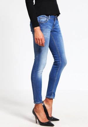 Vrouw gekleed in strakke blauwe spijkerbroek, zwart top met lange mouwen en zwarte puntige hoge hakken, staand tegen een witte achtergrond.