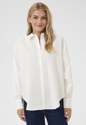 Witte button-up shirt van gladde stof, met een kraag, lange mouwen en een rechte zoom met zijsplitten. Gecombineerd met donkerblauwe jeans.