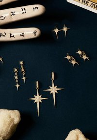 Gouden stervormige oorbellen en hangers versierd met sprankelende edelstenen, gerangschikt op een donkere ondergrond, met textuurstenen en een decoratief handmodel.