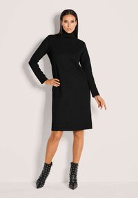 Frau in einem schwarzen langärmeligen Rollkragenkleid und genagelten schwarzen Knöchelstiefeln vor einem schlichten hellen Hintergrund.