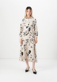 Femme debout portant une robe midi beige avec des imprimés floraux noirs et des escarpins noirs à bride arrière, sur un fond blanc uni.