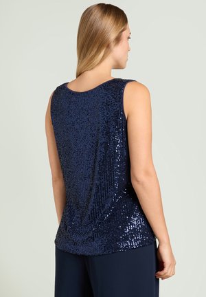 Frau mit blonden Haaren trägt ein ärmelloses, marineblaues Paillettenoberteil und passende Hosen, steht mit dem Rücken zur Kamera.