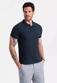 Camiseta tipo polo azul marino con mangas cortas, cuello acanalado, textura matizada y detalle de logo bordado en el pecho, combinada con pantalones de color gris claro.