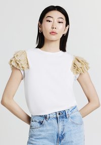 Camicia bianca a maniche corte con dettagli in rouches beige testurizzati sulle spalle, abbinata a pantaloncini in denim azzurro. Design semplice e vestibilità contemporanea.