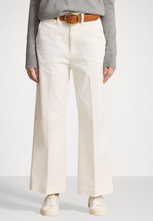 Pantalons larges en coton blanc avec plis à l'avant et taille haute, associés à une ceinture en cuir marron et des baskets blanches.