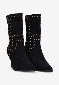 Botas de tobillo de ante negro con puntera afilada y tacón, adornadas con tachuelas doradas en un patrón decorativo a lo largo de los lados y las costuras.