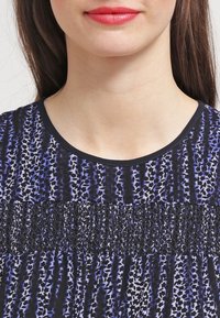 Blau und schwarz gemusterte Bluse mit Rundhalsausschnitt, mit strukturiertem Rüschen-Detail im Brustbereich und glattem Stoff.