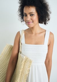 Balta, be rankovių, elastinė suknelė su kvadratiniu iškirpimu. Modelis laiko didelę, tekstūruotą, pintą šiaudų rankinę su nusidėvėjusiomis briaunomis.