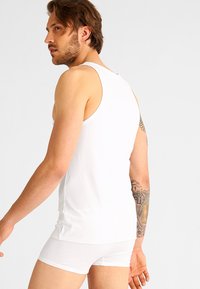 Un tricou alb din bumbac, fără mâneci, și șorturi boxer albe purtate de un model, evidențiind un design ajustat, o textură netedă și un tatuaj pe braț.
