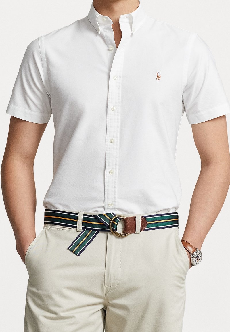 Man som bär en vit kortärmad skjorta med knappar, randig tygbälte med dubbel ringspänne, beige byxor och ett armbandsur med brun läderrem.