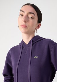 Violetinis džemperis su tekstūru, reguliuojamu raiščiu ir mažu žaliu logotipu ant krūtinės. Lengvas ir kasdienis dizainas.