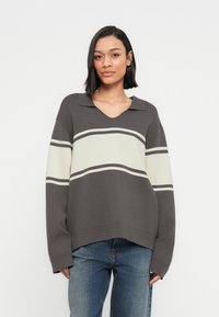 Γυναίκα με σκούρα, ίσια μαλλιά μέχρι τον ώμο, που φοράει γκρι και λευκό ριγέ πλεκτό πουλόβερ και μπλε τζιν, ποζάροντας μπροστά σε λευκό φόντο.