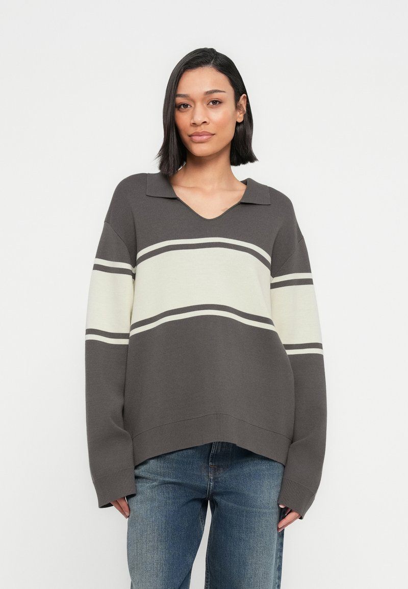 Γυναίκα με σκούρα, ίσια μαλλιά μέχρι τον ώμο, που φοράει γκρι και λευκό ριγέ πλεκτό πουλόβερ και μπλε τζιν, ποζάροντας μπροστά σε λευκό φόντο.