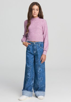 Giovane donna con lunghi capelli castani che indossa un maglione di paillettes rosa, jeans floreali azzurri a gamba larga e sneaker bianche, in piedi con una mano in tasca.