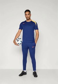 Kék sportpóló sárga díszítéssel és narancssárga logóval, kék nadrággal párosítva. A személy egy focilabdát tart, fekete sportcipőt visel.