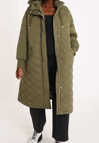 Olijfgroene gewatteerde lange jas met een capuchon, ritssluiting en zijzakken; heeft ribgebreide manchetten en een gestructureerde afwerking.