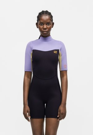 Femeie stând, privirea înainte, purtând un costum de scufundări cu mâneci scurte, negru și lavandă, cu sigle pe piept și mânecă.